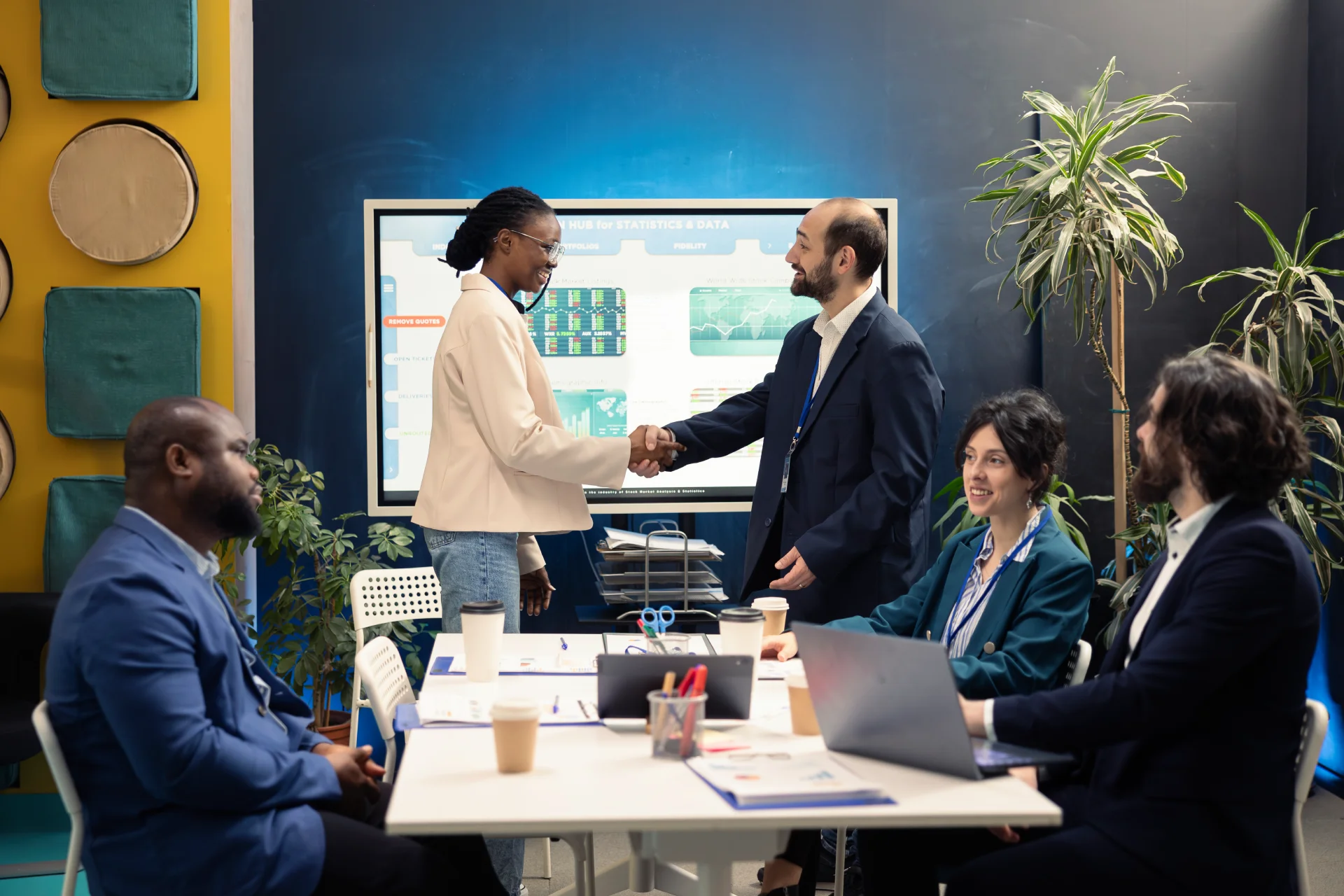Two professionals shaking hands during a business meeting while colleagues sit around a table with laptops and documents.