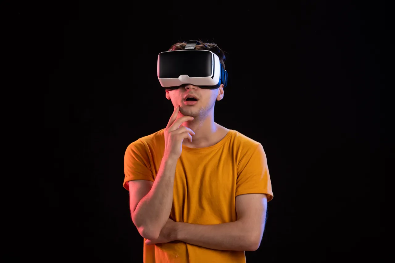 A smiling young man with glasses and curly hair, wearing a blue shirt, holding a silver laptop or tablet.